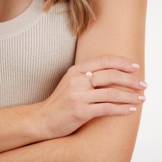 Close-up of a hand wearing a gold akoya pearl and diamond ring on a neutral background.